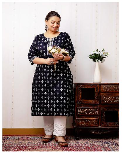 Black tunic with white pattern and trim at the neckline and sleeves, combined with white summer trousers -- photographed on a woman standing in an Indian-style room holding a bouquet of flowers.